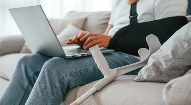 Ein männliche Person mit einer Schlinge um seinen Arm und einer Krücke neben ihm liegend, meldet auf einem Sofa mit seiner Unverletzten Hand einen Versicherungsfall.